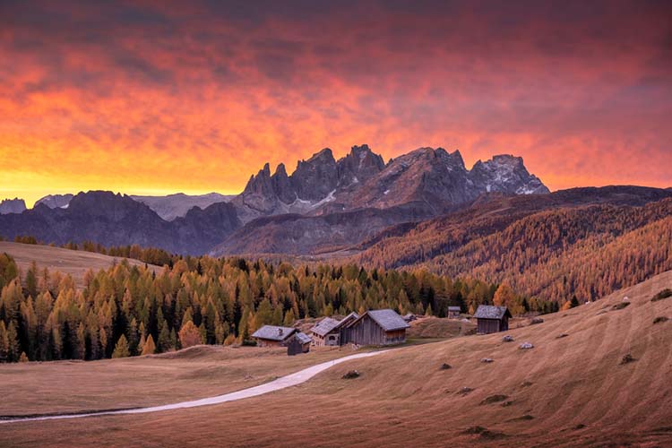 Beautiful red sunset at Fuchiade valley in Italian Dolomites countryside. Wooden huts, orange larches forest and snowy mountains peaks on background. Dolomite Alps, Italy