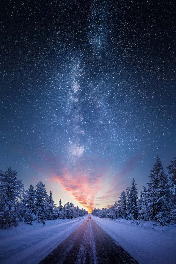 Road leading towards colorful sunrise between snow covered trees with epic milky way on the sky